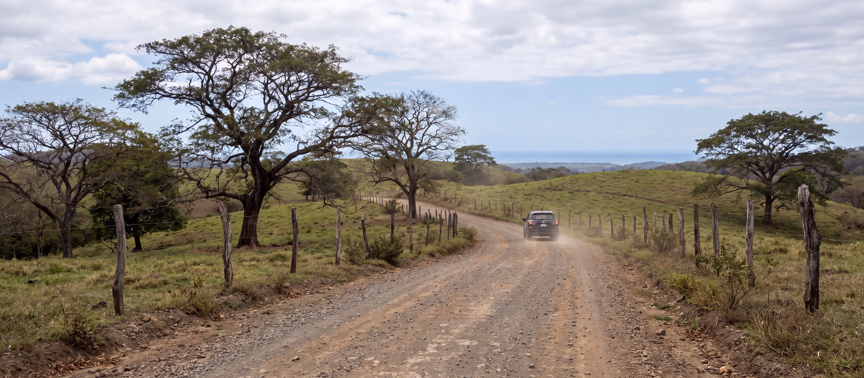 Erdpiste zwischen den Hügeln und Bäumen von Guanacaste, in der Ferne ein Geländewagen, am Horizont der Pazifik
