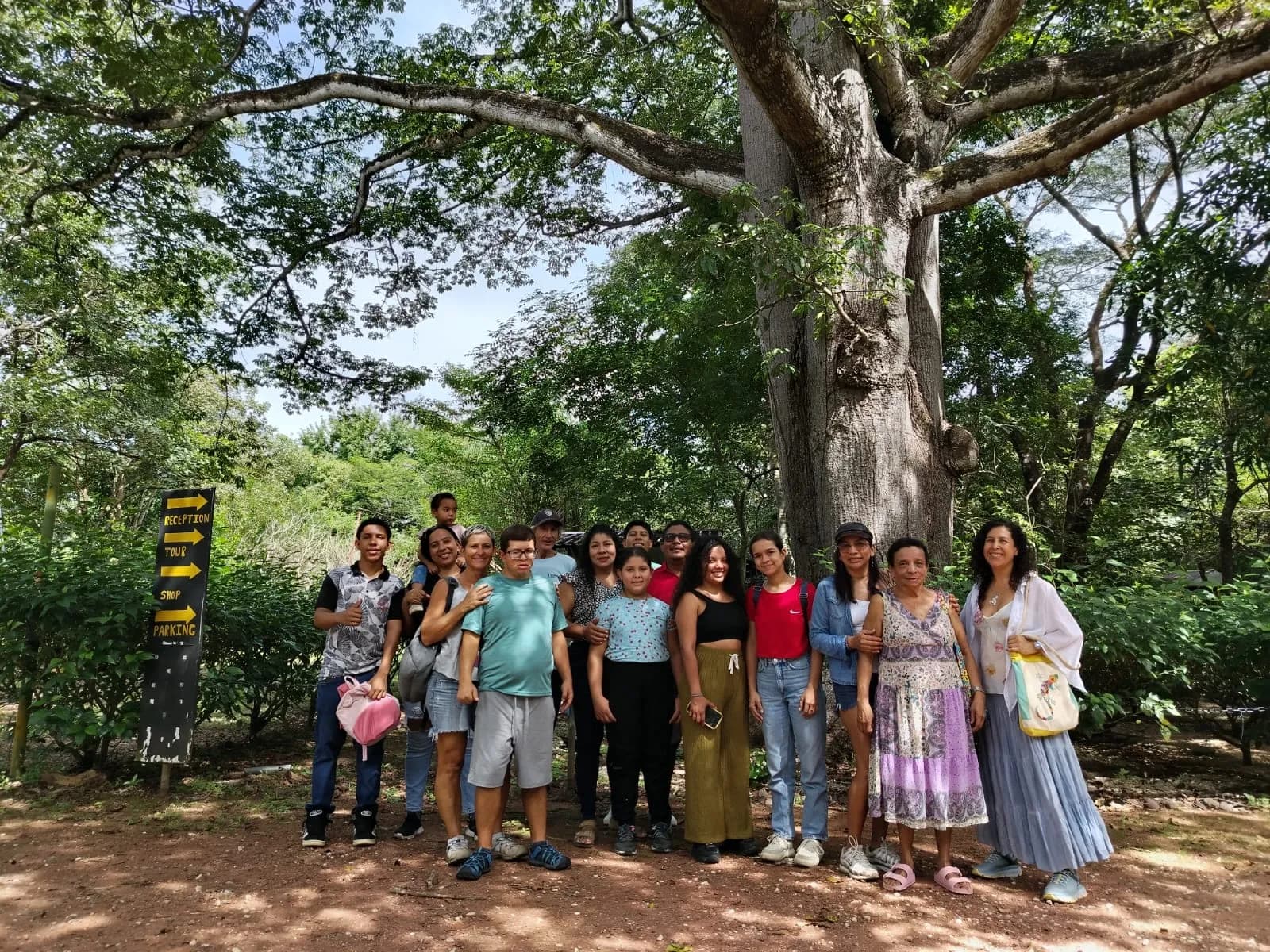 Groupe de visiteurs posant devant un immense ceiba, à côté d'un panneau indiquant l'accueil, la visite, la boutique et le parking